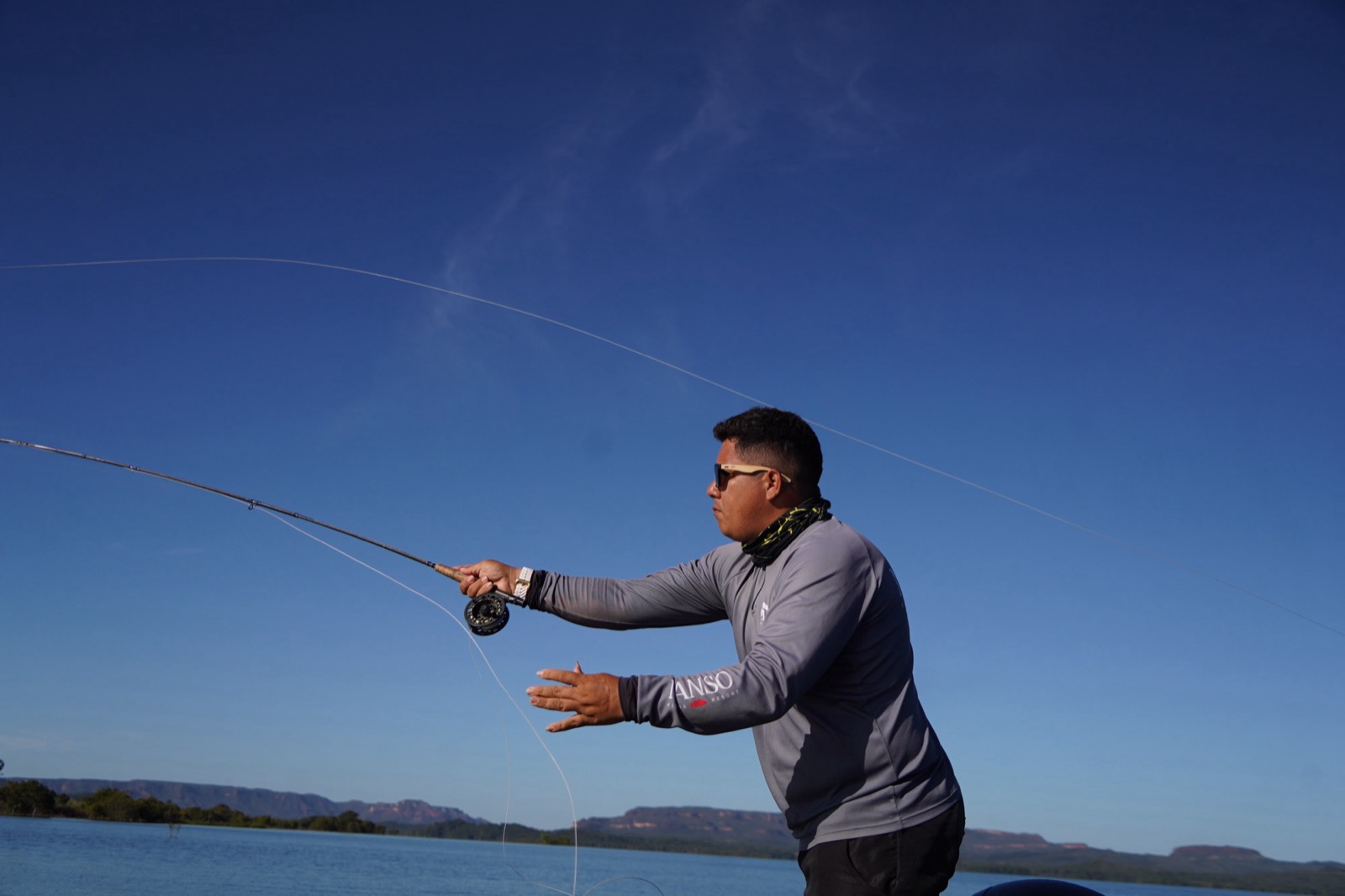 Pescador em ação de fly fishing com céu azul e serra ao fundo no Lago do Manso